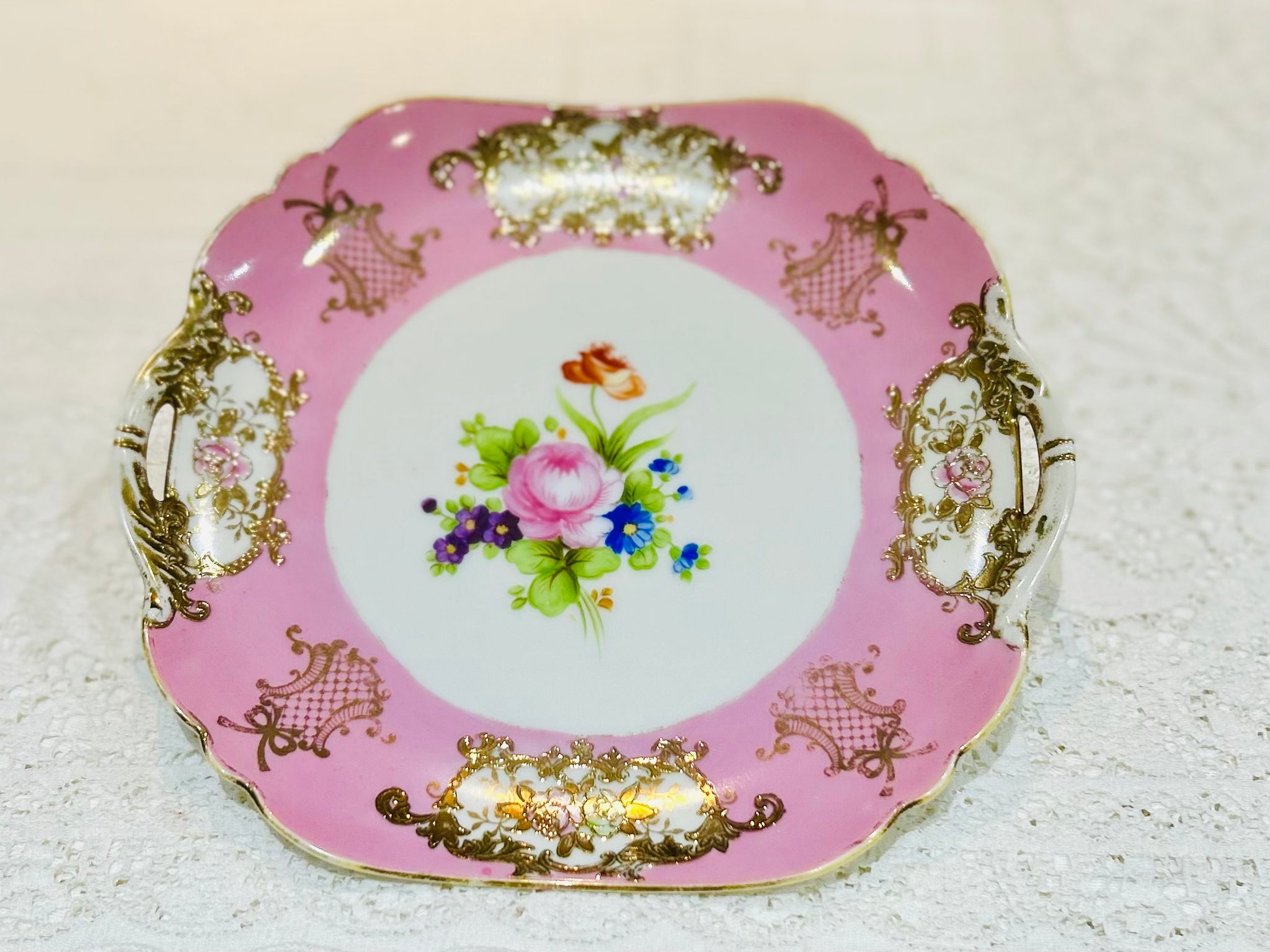An elegant and lavishly detailed plate made by Noritake China.  The outer banding is a bright candyfloss pink. The inner circle is white with a painted display of pink cabbage roses and flowers.  Overall there is no damage, some rubbing to the gilding.  The width is 9 1/2".  Side handles.  Makers stamp to base.